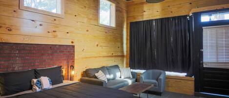 A wooden cabin room with a bed, two sofas, a table, and a window with curtains.