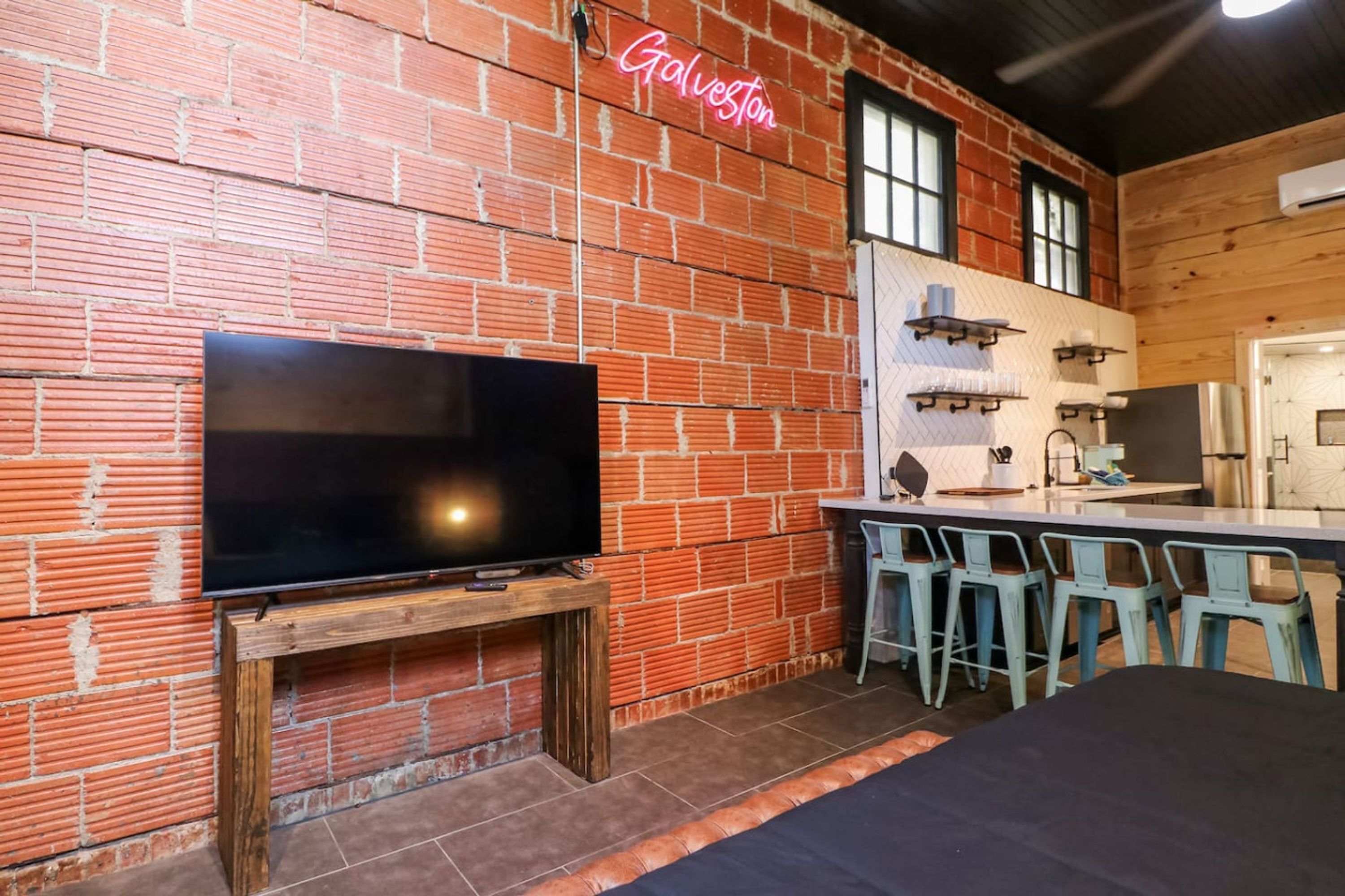 Una stanza con pareti in mattoni, una TV a schermo piatto montata su un supporto in legno, un neon con scritto 'Galveston' e un'area bar con sgabelli.