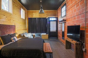 A wooden cabin-style bedroom with a bed, two chairs, a bench, a TV, and a window with curtains. - Restored Historic Loft - 3 Mins to the Beach (Galveston)