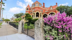 Ein großes, rot-ziegel-Mansion mit Rundbogenfenstern und einem Turm, umgeben von einem schwarzen Metallzaun und üppiger Bepflanzung.