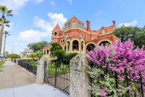 A large, red-brick mansion with arched windows and a turret, surrounded by a black metal fence and lush greenery. - Restored Historic Loft - 3 Mins to the Beach (Galveston)
