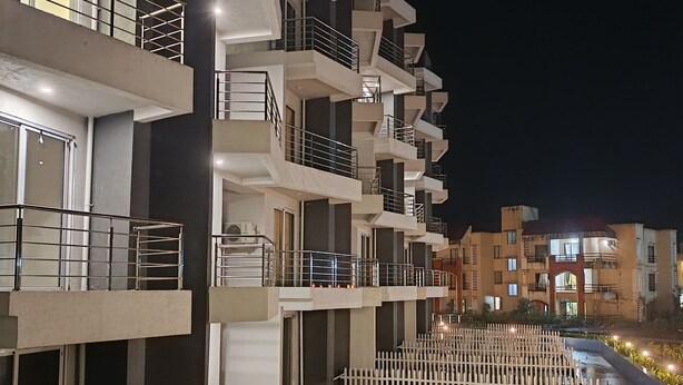 A multi-story building with balconies, a white picket fence, and a green lawn. - Karjat Poolside Studio | Pvt Balcony | Couples, Friends & Family | Near Mumbai (Karjat)