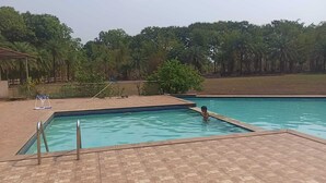 A rectangular swimming pool with a tiled deck, surrounded by trees and a fenced area. - Karjat Poolside Studio | Pvt Balcony | Couples, Friends & Family | Near Mumbai (Karjat)