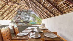 Un coin repas avec une table en bois, dressée pour quatre personnes, entourée de chaises, le tout sous un toit de chaume.
