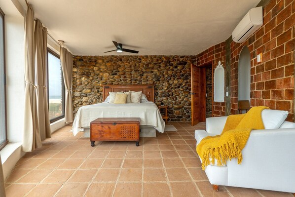 Un dormitorio con pared de piedra, cama con ropa blanca, cómoda de madera, sofá blanco con una manta amarilla y un ventilador de techo.