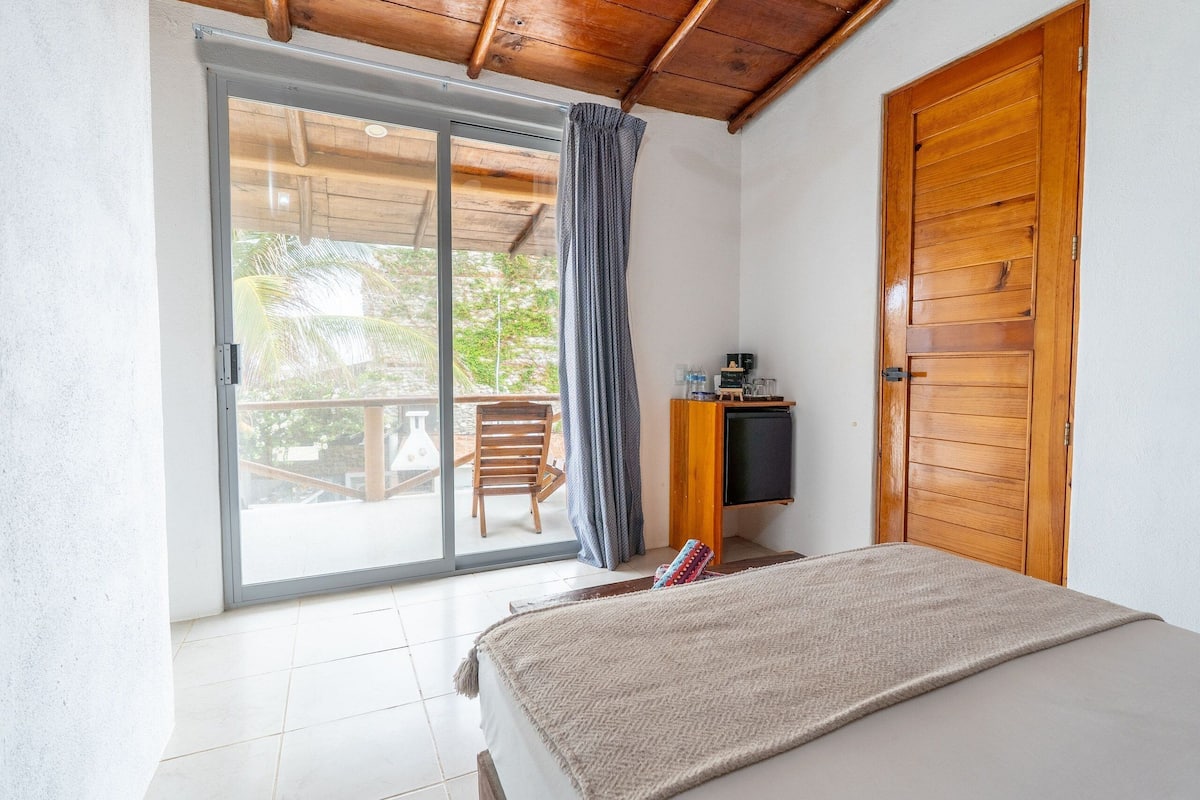 A bedroom with a bed, a wooden door, a TV, and a sliding glass door leading to an outdoor area with a chair and table.