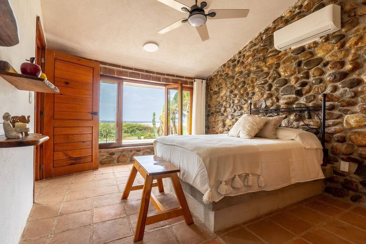 A bedroom with a stone wall, a bed with white bedding, a wooden stool, a ceiling fan, and a window with a view of greenery.