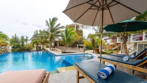Un espace aménagé au bord de la piscine, avec des chaises longues, un grand parasol et une vue sur un bâtiment doté d’une piscine.