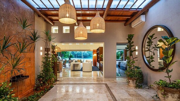 A spacious living area with a high ceiling, large windows, and a view of the outdoors. There are multiple hanging lights and a variety of plants.