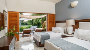 A hotel room with two beds, a sitting area with a table and chairs, and a view of a tropical garden.