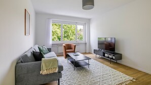 A modern living room with a grey sofa, a small table, a TV stand, and a window with a view of trees. - Blueground | Basel-Vorstaedte, view, nr Rhine (Basel)