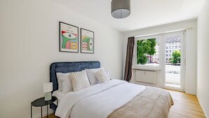 A bedroom with a bed, a nightstand, a lamp, two framed artworks, a window with curtains, and a view of buildings outside. - Blueground | Basel-Vorstaedte, view, nr Rhine (Basel)