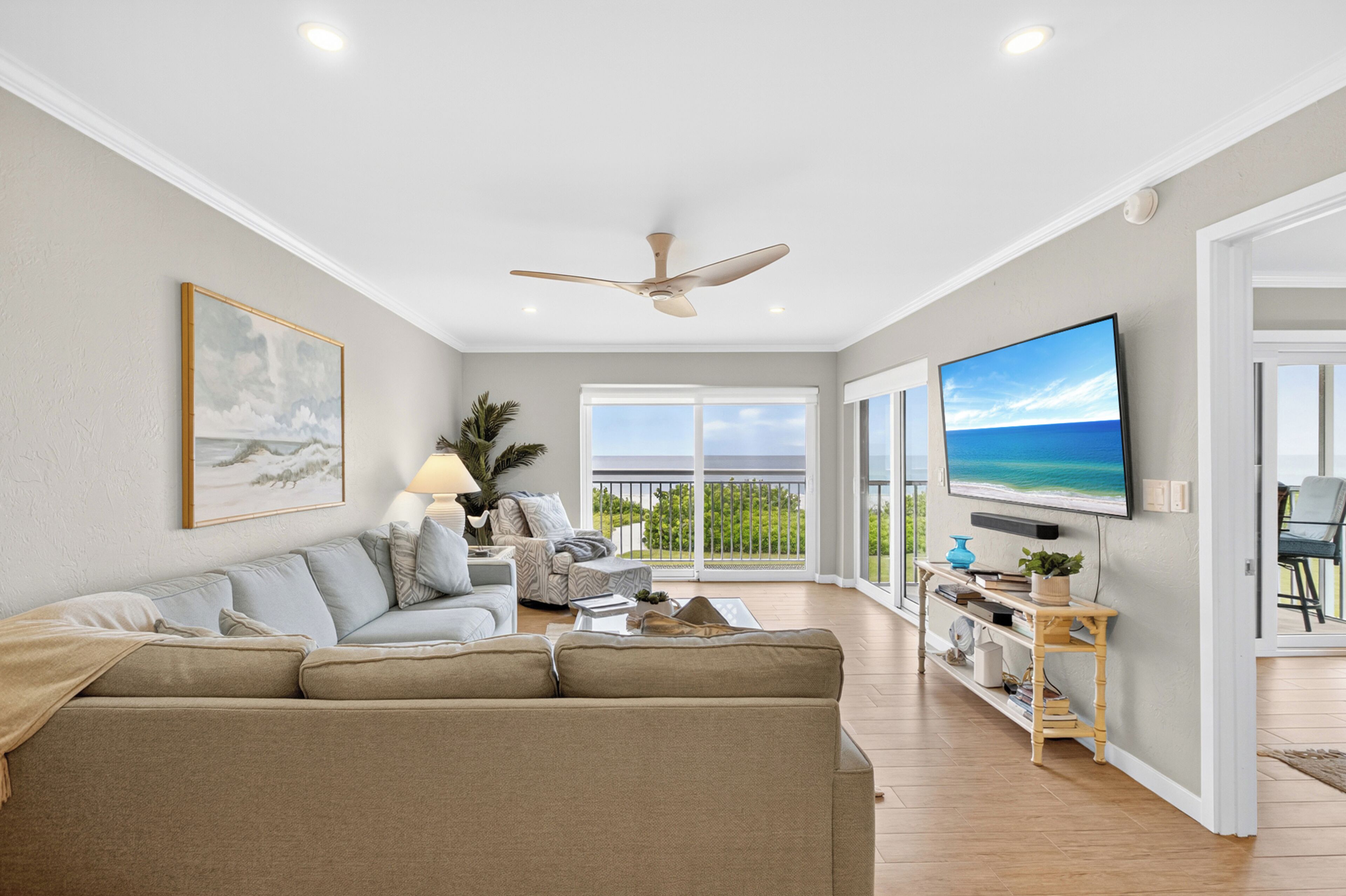 A spacious living room with a large flat-screen TV, a ceiling fan, and a balcony with a view.