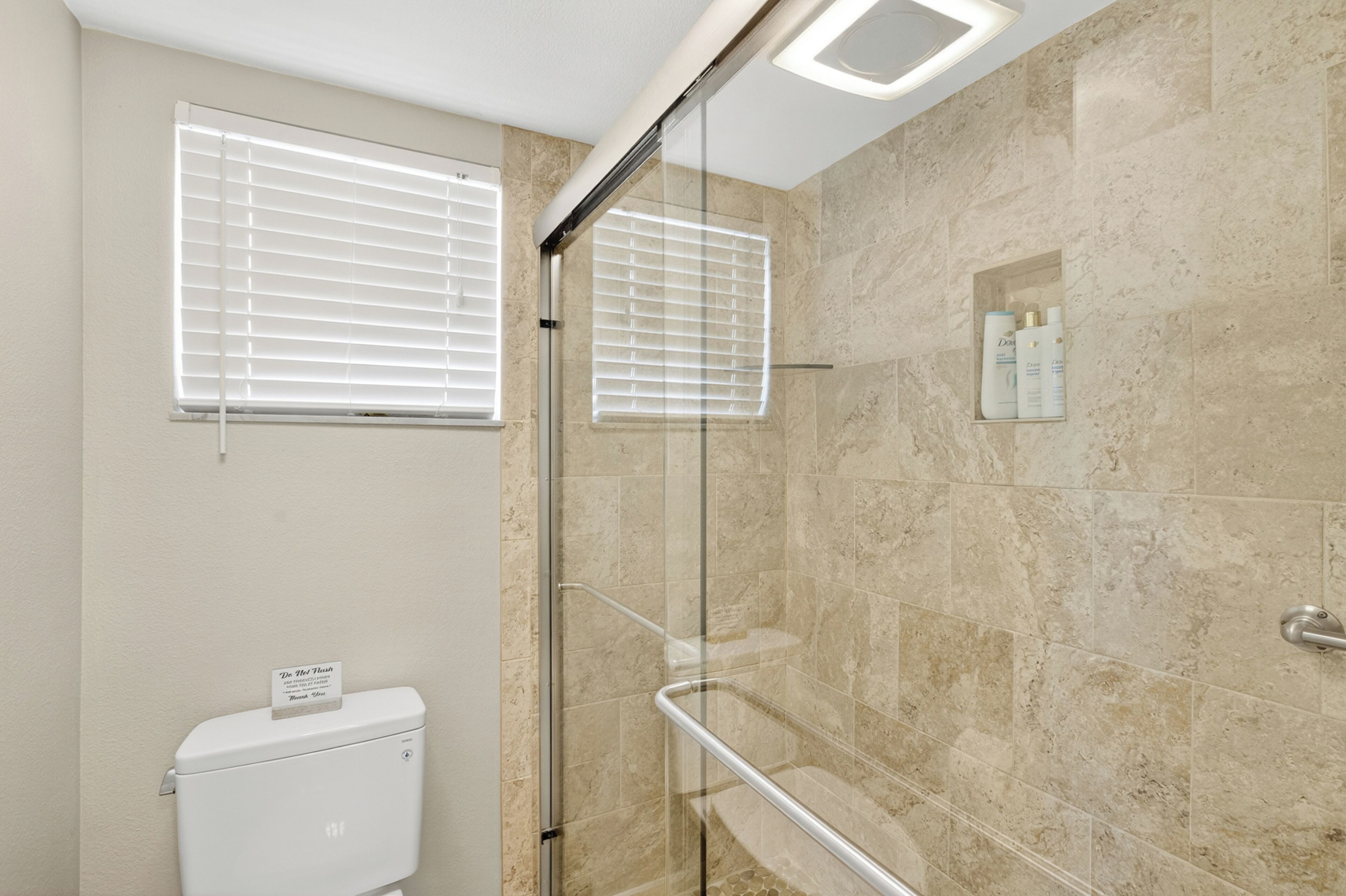 A modern bathroom with a glass shower enclosure, beige tiled walls, and a toilet with a paper towel dispenser.