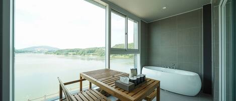 Une salle de bain moderne avec une grande baignoire, une table en bois avec un plateau, et une vue sur un lac grâce à de grandes fenêtres.