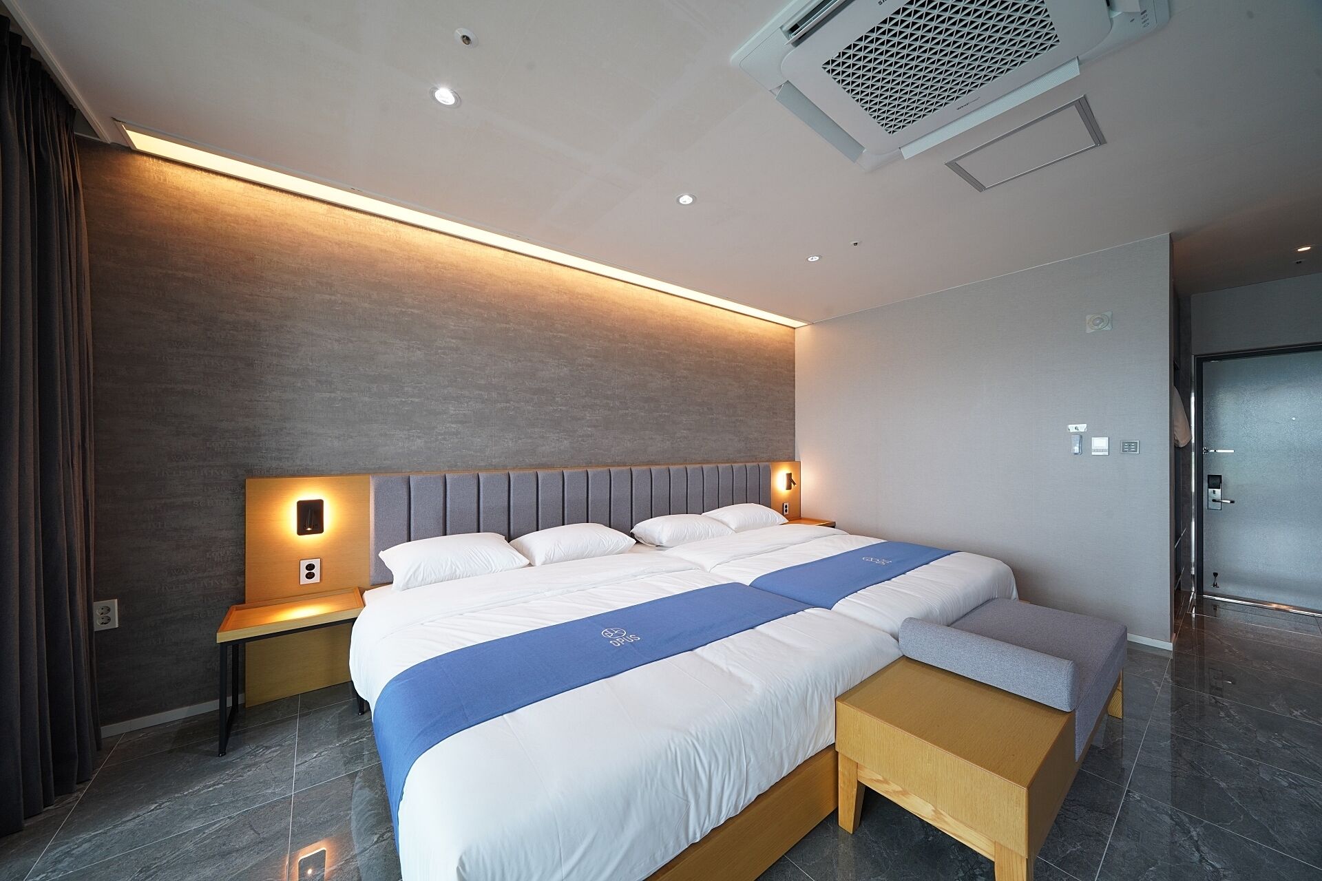 Une chambre d’hôtel moderne, dotée d’un grand lit, de tables de chevet, d’un banc et d’un climatiseur fixé au mur.