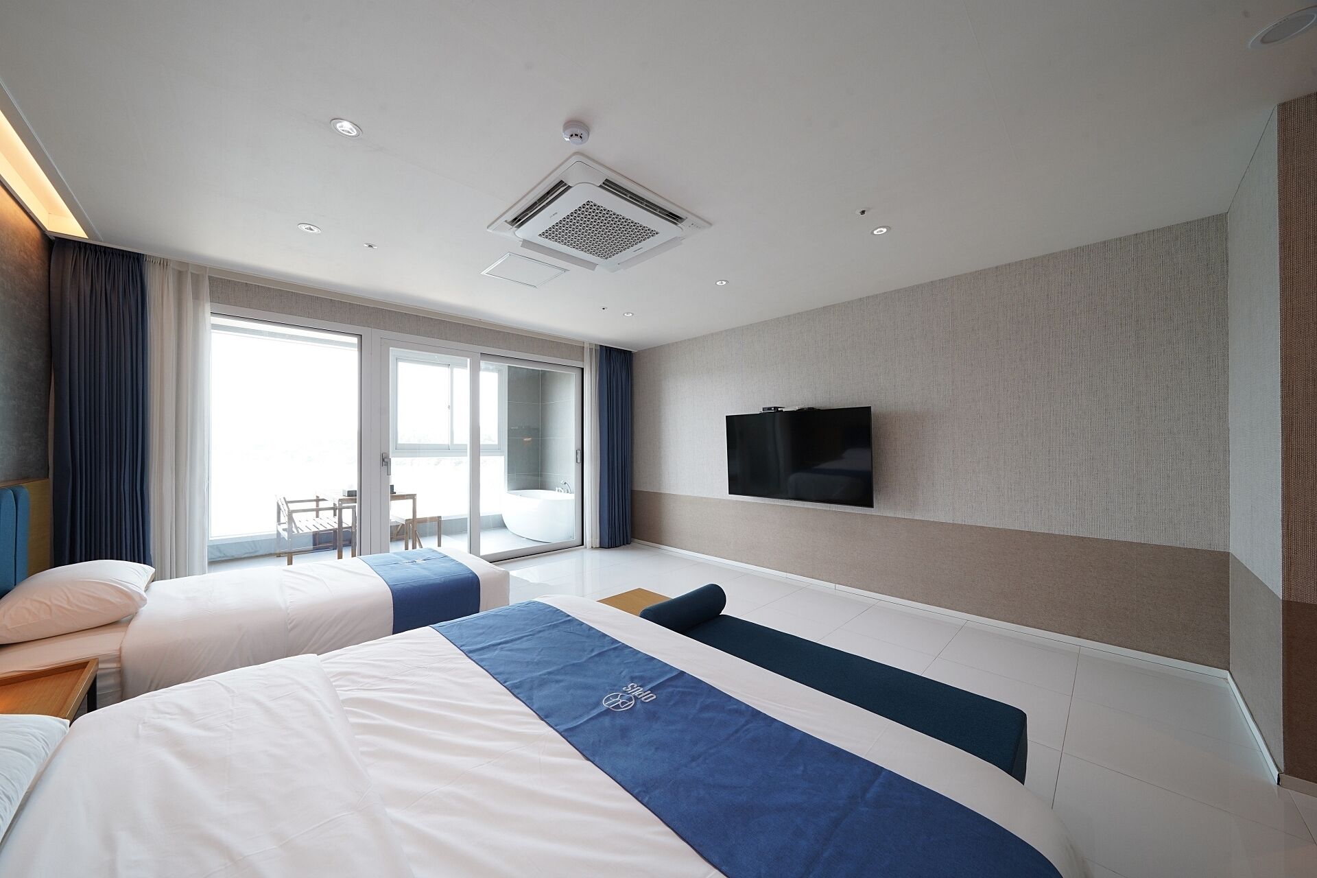 A hotel room with two beds, a large window with curtains, a flat-screen TV mounted on the wall, and a ceiling air vent.