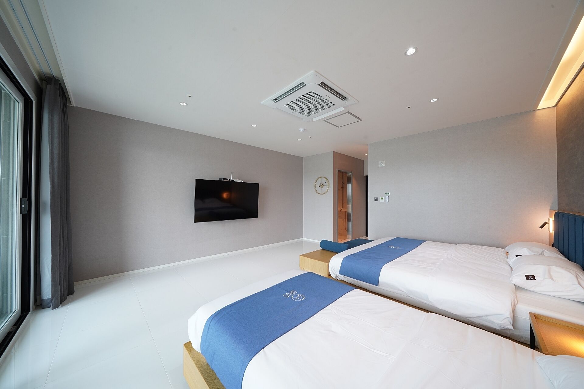 A modern hotel room with two beds, a flat-screen TV mounted on the wall, a ceiling vent, and a door leading to another room.