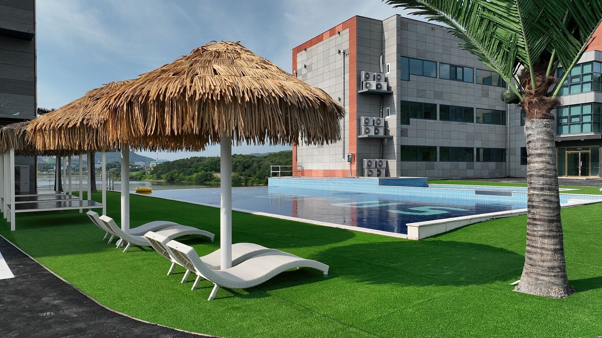 A modern pool area with artificial grass, lounge chairs, and a thatched umbrella.