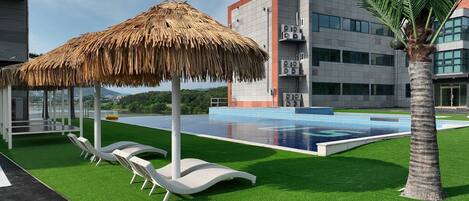 A modern pool area with artificial grass, lounge chairs, and a thatched umbrella.