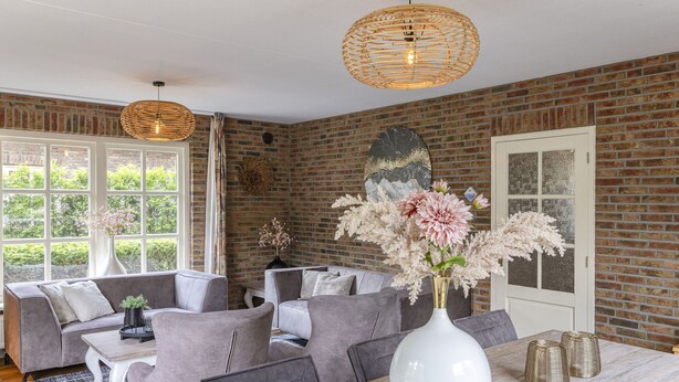 A modern living room with a brick wall, grey sofas, a white dining table, and a large window.