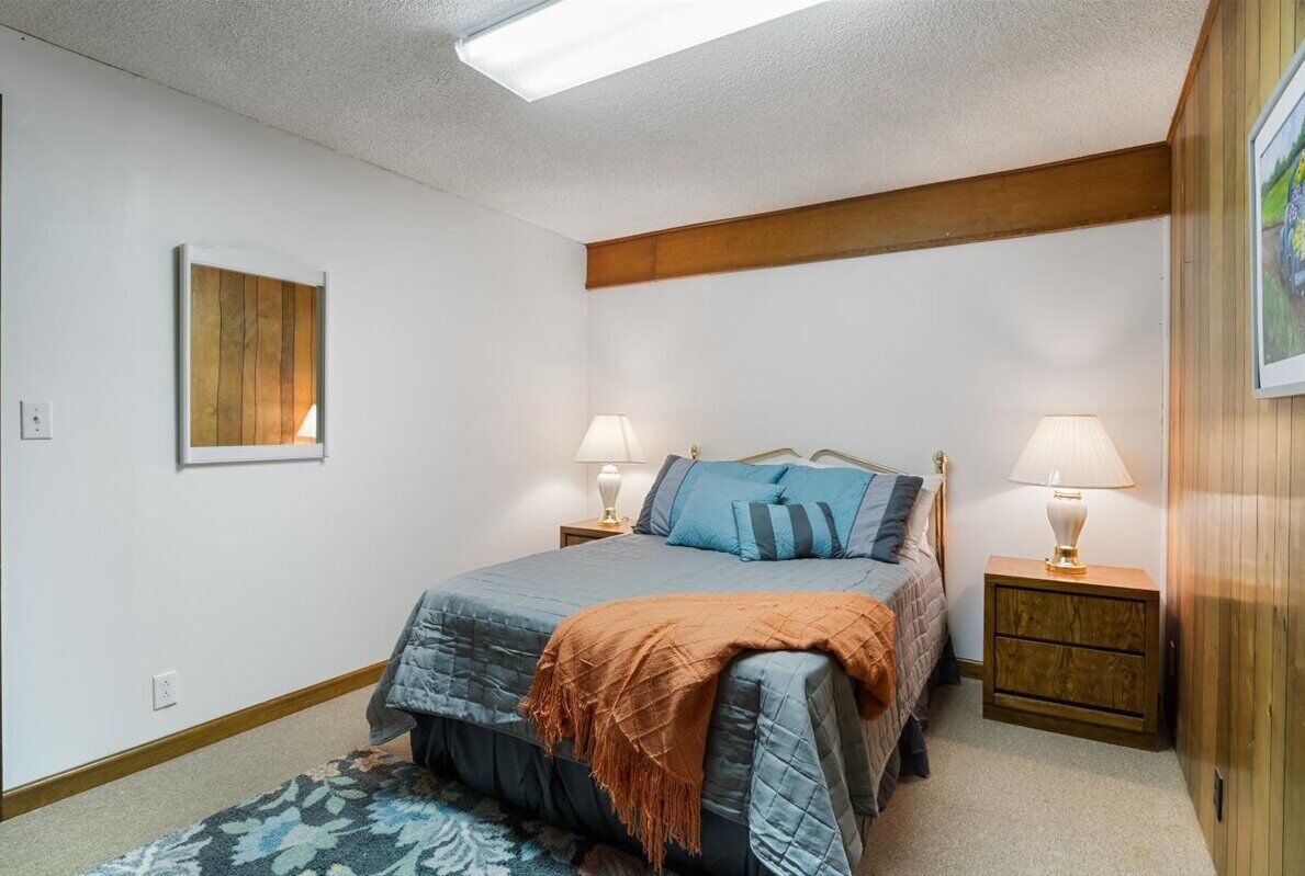 Un dormitorio con cama, dos mesitas de noche con lámparas, cabecera de madera, un cuadro enmarcado en la pared y una alfombra estampada.