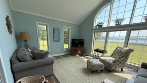A living room with a grey sofa, a patterned armchair, a television, and a large window overlooking a lake.