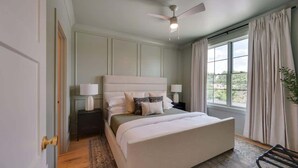Un dormitorio con una cama grande, un ventilador de techo, una mesita de noche, una ventana con cortinas y vistas al exterior.