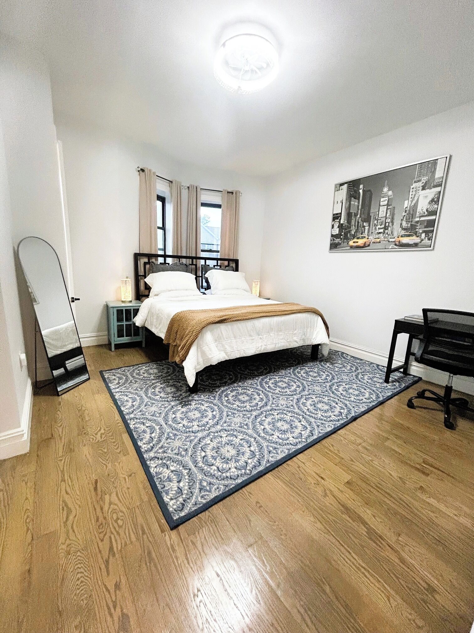 Un dormitorio con cama, escritorio, silla, espejo, ventana con cortinas e imagen colgada en la pared.