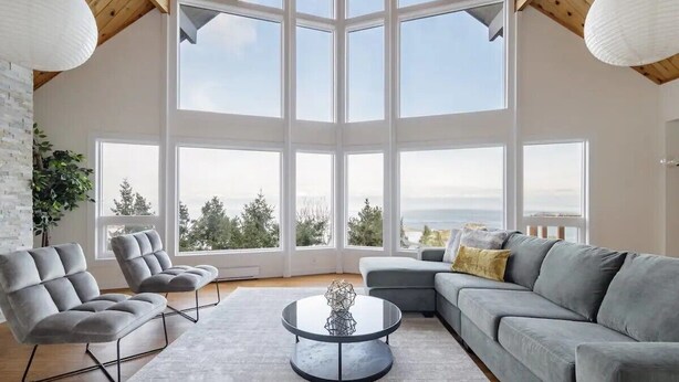 A spacious living room with a high ceiling, large windows offering a view of the outdoors, and modern furniture. - Ocean's Embrance Suite (Nanaimo)