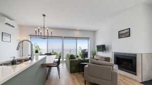 A modern kitchen with a dining area, a fireplace, and a large window overlooking the sea.