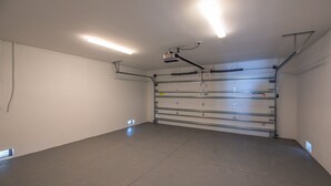 Un garage propre et dégagé, éclairé par des néons, avec une porte de garage en métal et des tuyaux apparents au plafond.
