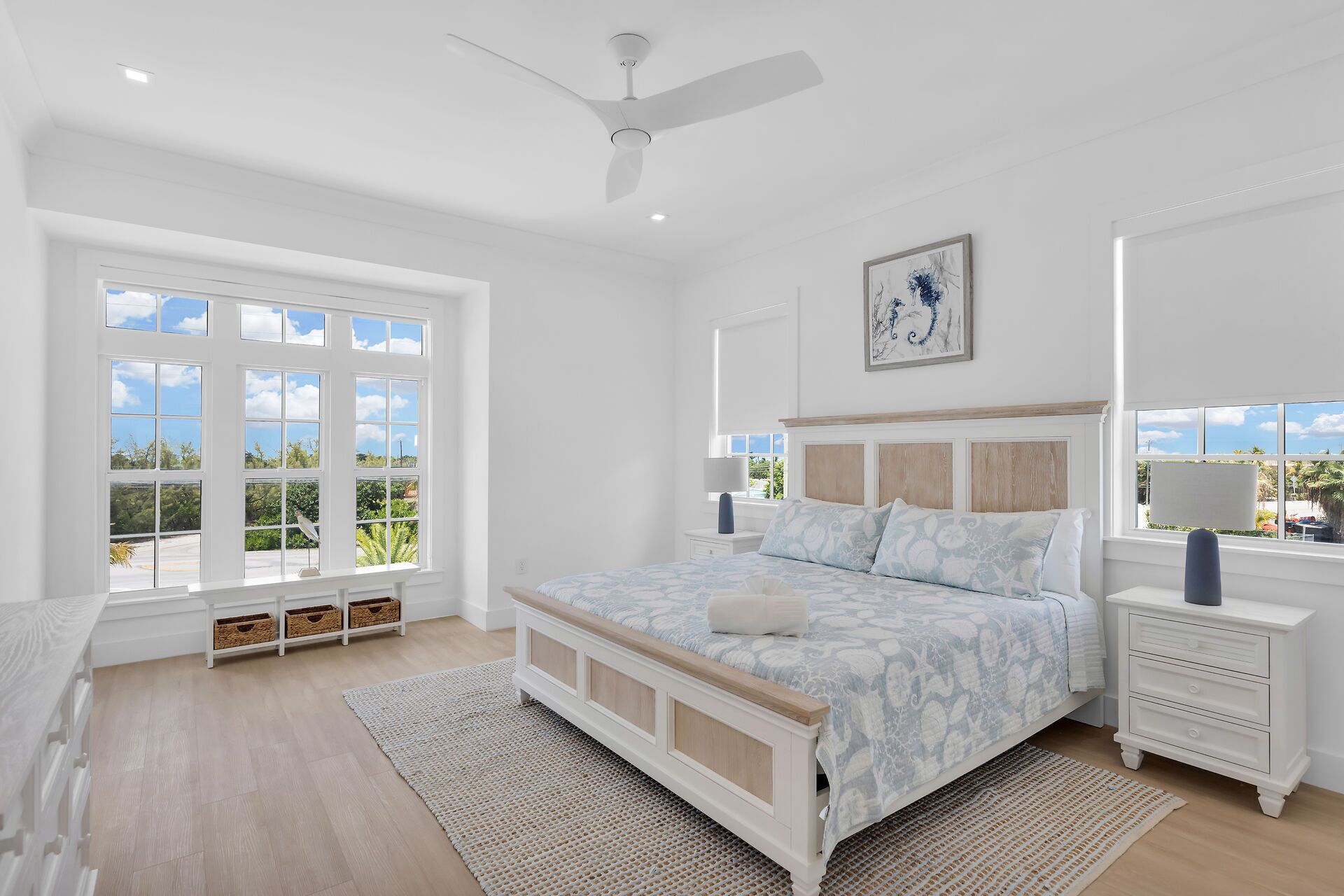 A bedroom with a large bed, a ceiling fan, and a view of greenery through the windows.