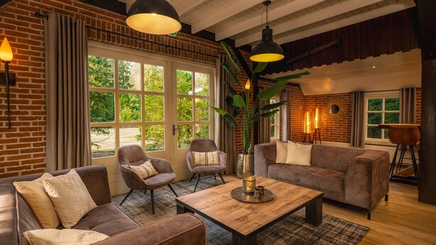 Een moderne woonkamer met een bakstenen muur, grote ramen en een gezellige zithoek met banken en stoelen.