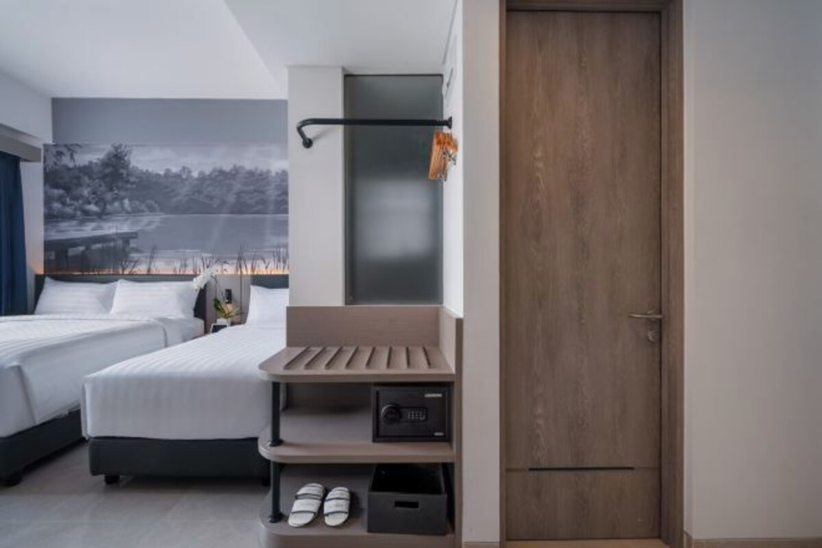 Una habitación de hotel moderna con dos camas, un pequeño espacio de descanso, un mueble para guardar zapatos y una puerta.