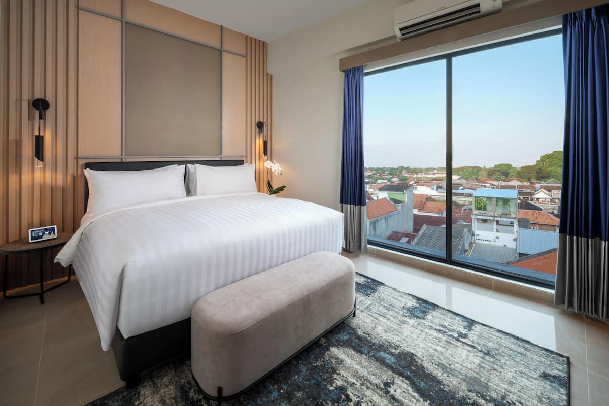 A modern hotel room with a large bed, a bench, a nightstand, a wall-mounted air conditioner, and a large window with a city view.