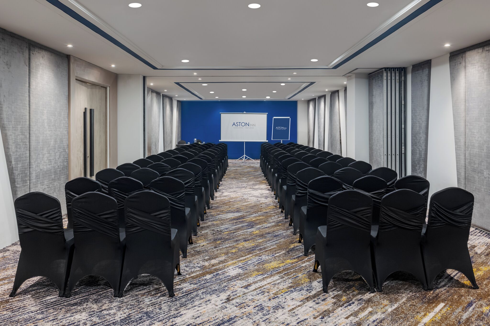 A conference room with rows of black chairs, a blue carpet, and a projection screen displaying "ASTON NNN."