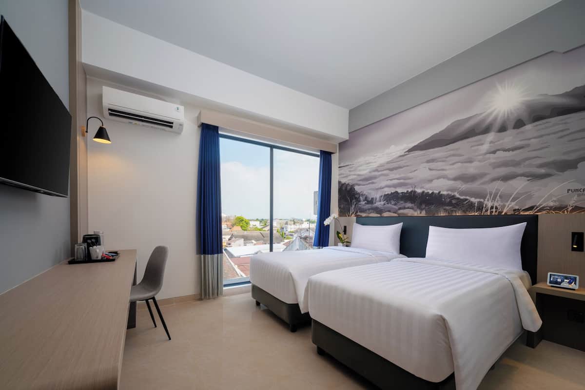 A hotel room with two beds, a desk, a chair, a TV, and a large window with a view of the city.