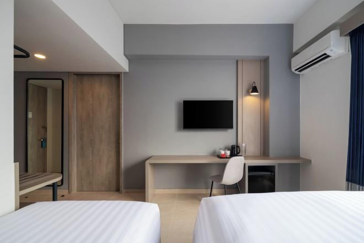 A hotel room with two beds, a desk, a TV, and an air conditioning unit.
