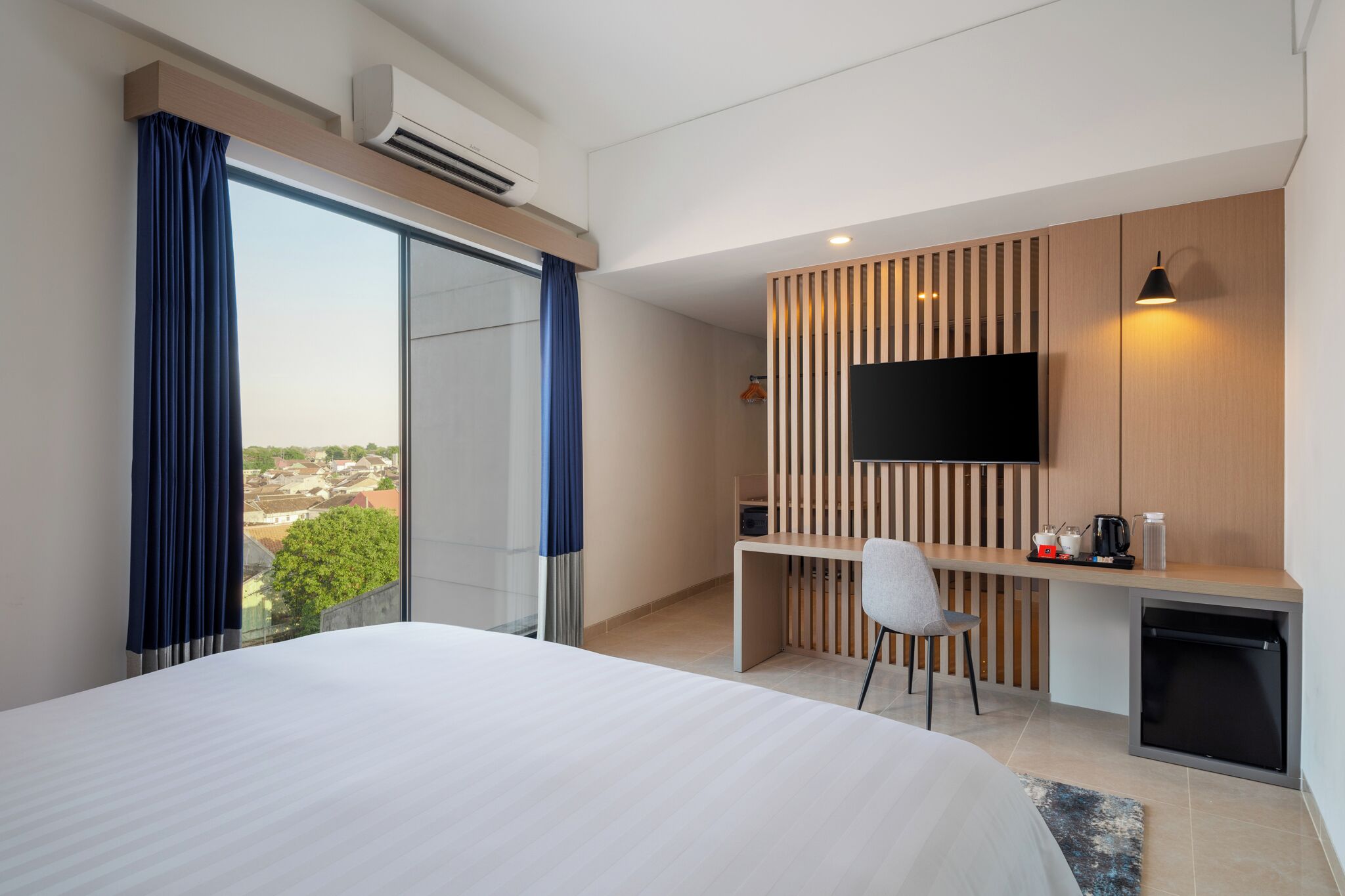 Ein modernes Hotelzimmer mit einem großen Bett, einem Schreibtisch mit Stuhl, einem Flachbildfernseher und Blick auf die Stadt durch das Fenster.