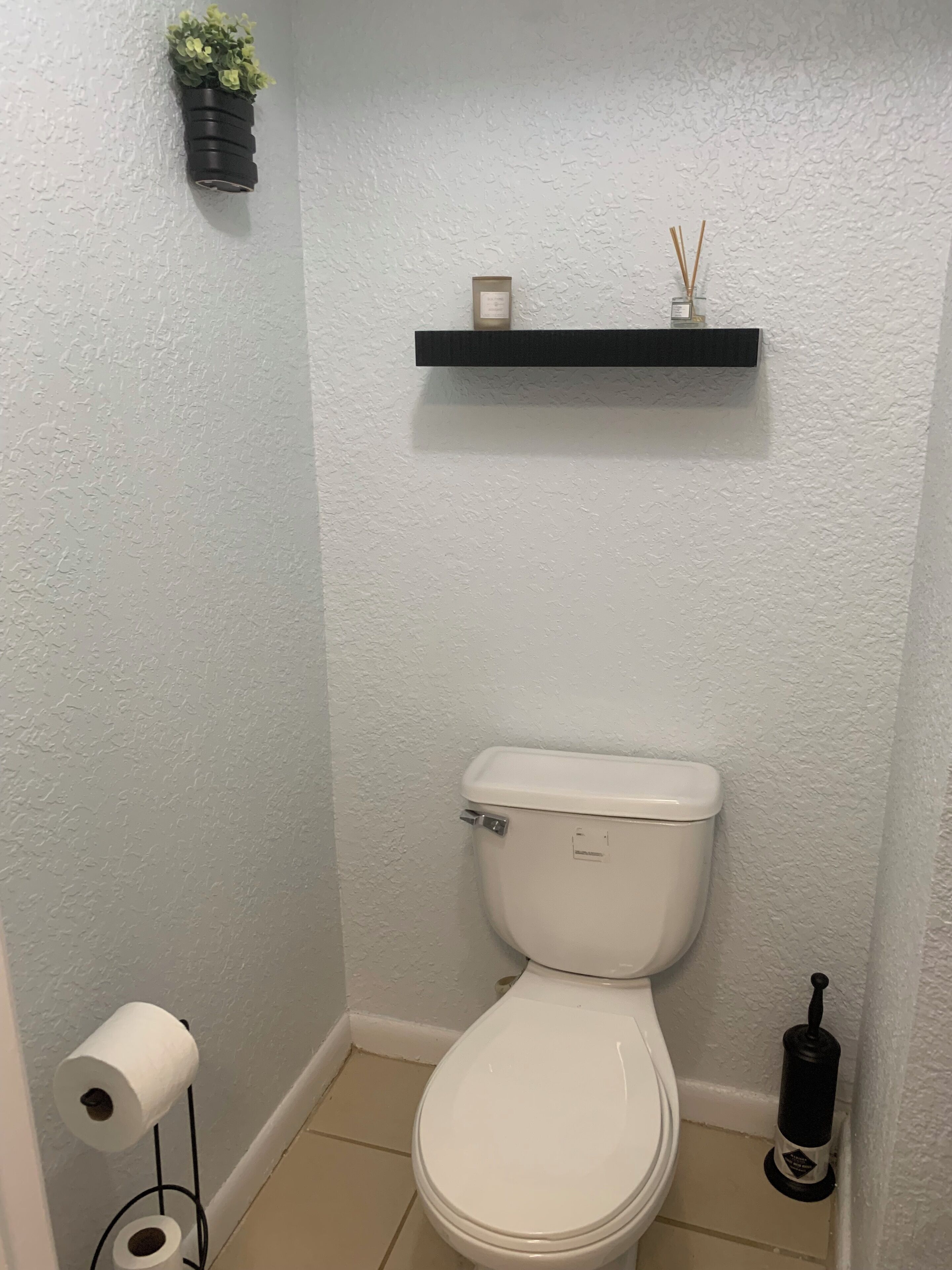 Casa de banho pequena com sanita, uma estante com uma planta e uma vela, e um suporte para papel higiénico.