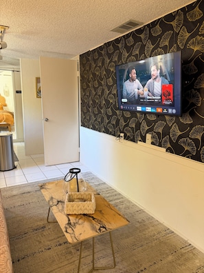 Une pièce avec un téléviseur fixé au mur diffusant un film, une table en bois sur laquelle se trouve un récipient en verre, et un mur orné d’un motif.