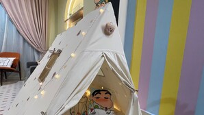 Una estructura tipo tienda de campaña con un personaje de caricatura en su interior, frente a una pared a rayas.