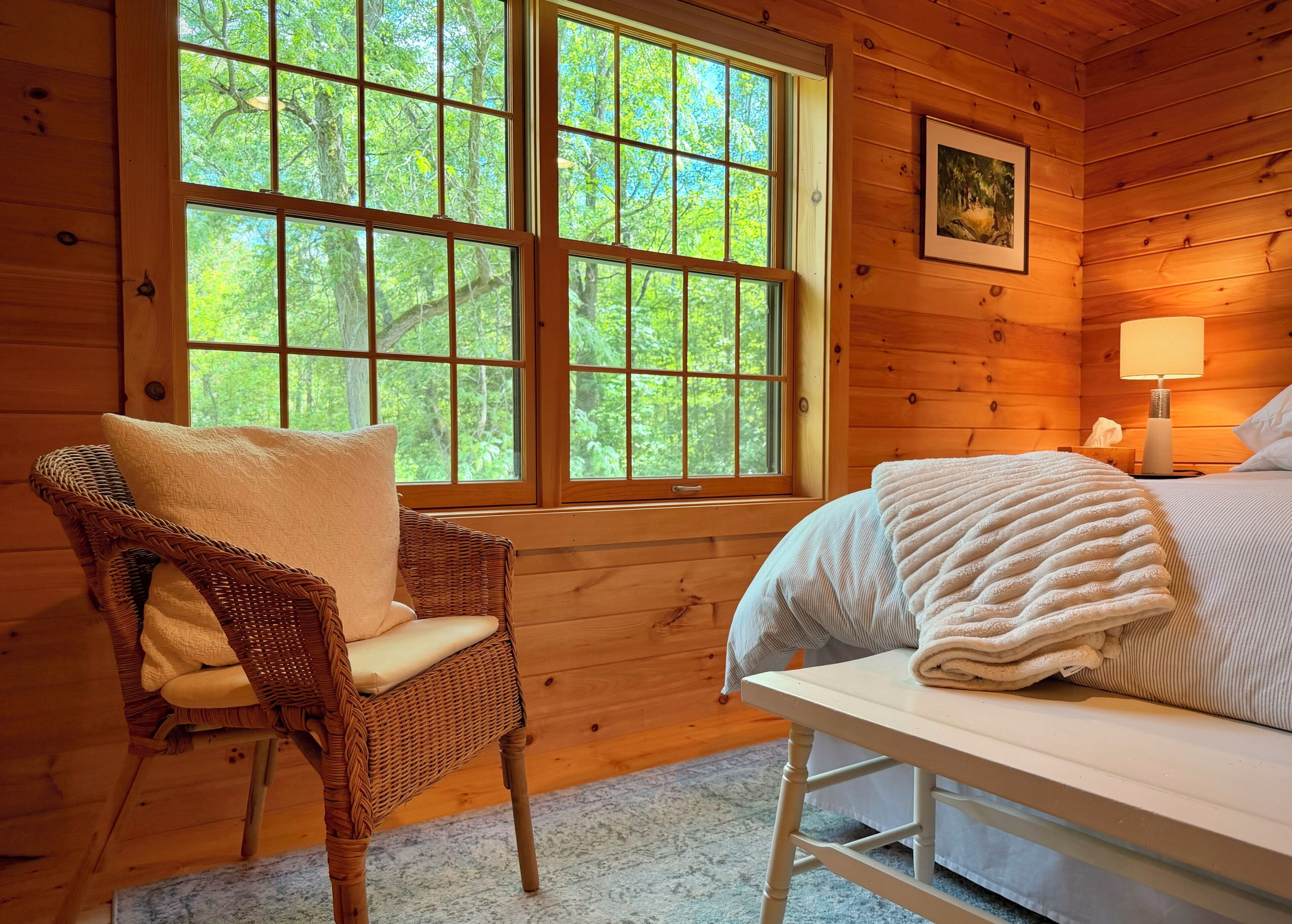 Habitación de cabaña de madera con una cama, una silla, un ventanal con vistas a árboles y un cuadro en la pared.