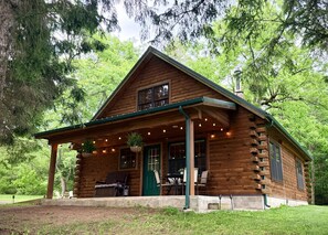 A log cabin with a porch, outdoor seating, and hanging plants. - Timber Haven-Modern cabin retreat with fireplace, AC, fire pit, grill, WiFi (Cohocton)