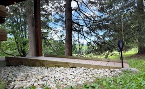 A wooden deck with a metal railing, surrounded by a gravel area and greenery. - Timber Haven-Modern cabin retreat with fireplace, AC, fire pit, grill, WiFi (Cohocton)