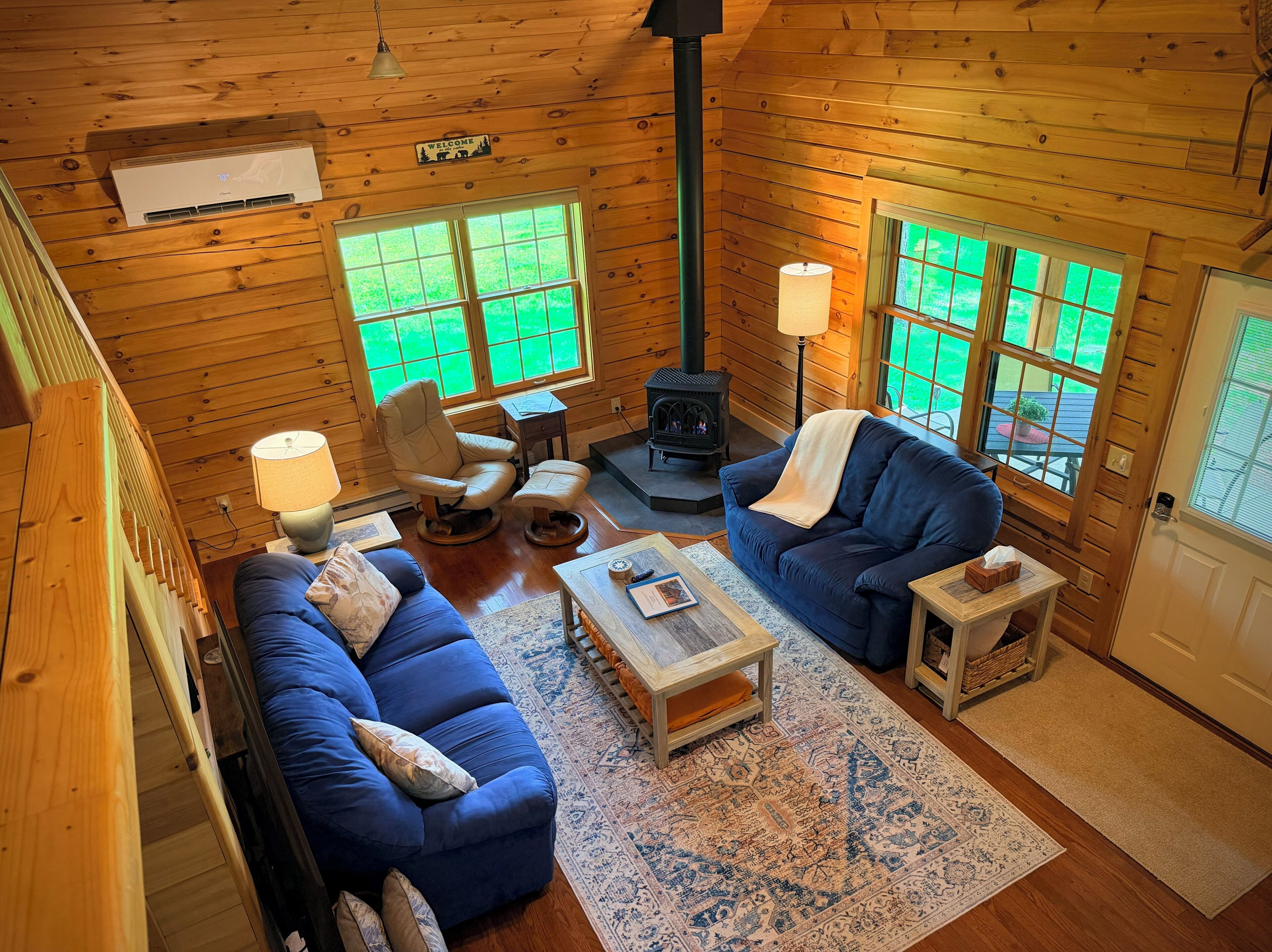 A cozy cabin interior with a sofa, armchair, and a small table, a fireplace, and large windows.
