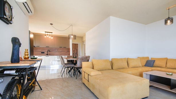 Un salon moderne avec un grand canapé d’angle beige, un coin repas comprenant une table et des chaises en bois, et une cuisine en arrière-plan.
