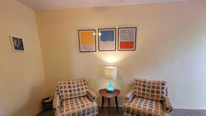 Uma sala de estar com dois poltronas, uma mesa redonda pequena com um abajur e três obras de arte emolduradas na parede.