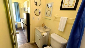 Un baño pequeño con inodoro, lavabo y dos espejos. Una toalla cuelga en un perchero, y hay un estante de vidrio sobre el lavabo.